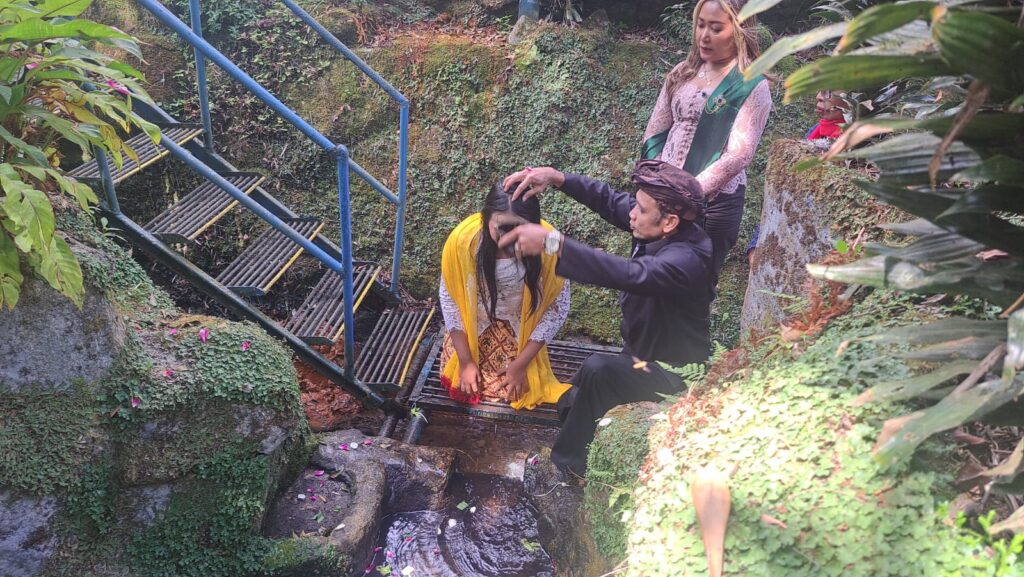 A man in a dark shirt blesses or places his hands on a woman's head in a yellow shawl, while she bows; a woman in a green sash watches and other onlookers stand nearby on a mossy stone stairway beside a small stream in an outdoor setting.